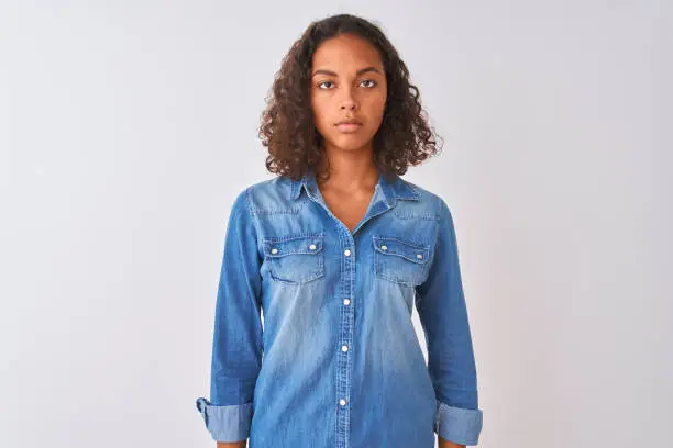 Photo of Young brazilian woman wearing denim shirt standing over isolated white background Relaxed with serious expression on face. Simple and natural looking at the camera.