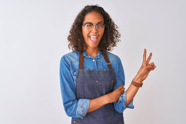 エプロンとメガネをかけた若いブラジル人シェフの女性が、勝利のサインをしているカメラで幸せそうな顔をして微笑む。2番 - baker occupation ストックフォトと画像