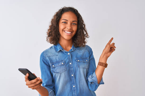 joven mujer brasileña usando teléfono inteligente de pie sobre fondo blanco aislado muy feliz señalando con la mano y el dedo hacia un lado - teenage girls cheerful smiling one person fotografías e imágenes de stock