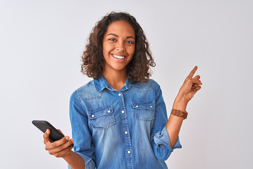 Young brazilian woman using smartphone standing over isolated white background very happy pointing with hand and finger to the side