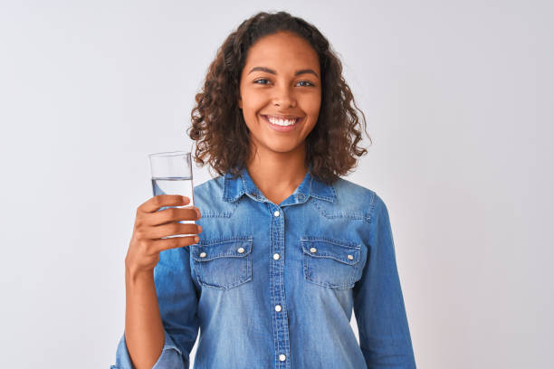 junge brasilianische frau hält glas wasser stehend über isolierten weißen hintergrund mit einem glücklichen gesicht stehen und lächelnd mit einem selbstbewussten lächeln zeigt zähne - toothy smile relaxation white healthcare and medicine stock-fotos und bilder