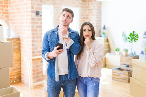 Beautiful young couple moving to a new house using smartphone serious face thinking about question, very confused idea Beautiful young couple moving to a new house using smartphone serious face thinking about question, very confused idea two people thinking stock pictures, royalty-free photos & images