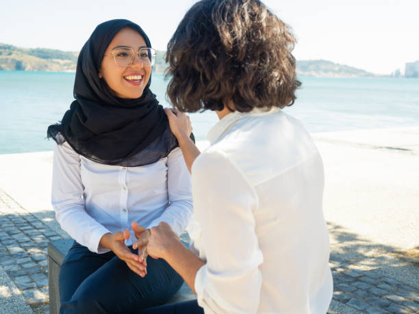 Multicultural female colleagues chatting and laughing outside during work break. Woman in hijab sharing good news with her coworker. Corporate friendship concept