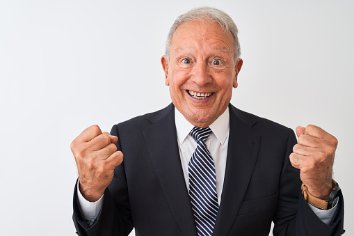 Senior grey-haired businessman wearing suit standing over isolated white background celebrating surprised and amazed for success with arms raised and open eyes. Winner concept.