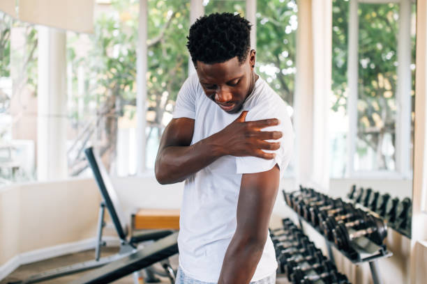 Sportsman holding painful shoulder in gym Side view of muscular African American man standing and suffering from shoulder pain during workout with dumbbells. Sportsman holding sore shoulder in gym black male massage stock pictures, royalty-free photos & images