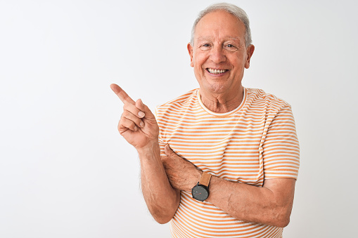 Hombre de pelo gris senior con camiseta a rayas de pie sobre fondo blanco aislado con una gran sonrisa en la cara, apuntando con el dedo de la mano hacia el lado mirando a la cámara. photo