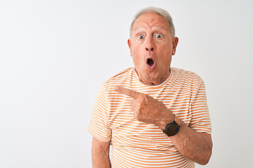 Senior grey-haired man wearing striped t-shirt standing over isolated white background Surprised pointing with finger to the side, open mouth amazed expression.