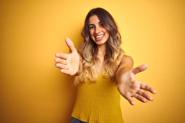 junge schöne frau trägt t-shirt über gelb isolierten hintergrund blickend auf die kamera lächelnd mit offenen armen für umarmung. fröhlicher ausdruck, der glück umarmt. - spanish and portuguese ethnicity horizontal looking at camera indoors stock-fotos und bilder