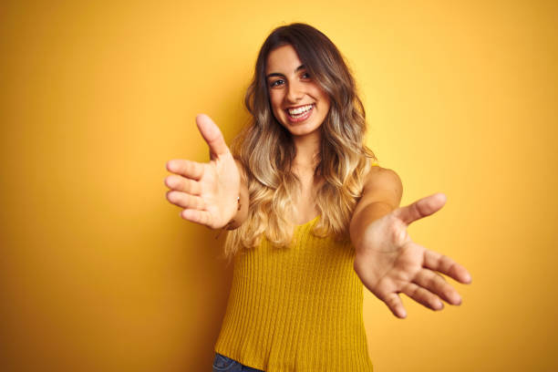 молодая красивая женщина в футболке на желтом изолированном фоне смотрит на камеру, улыбаясь с открытыми объятиями для объятий. веселое вы� - expressive hands стоковые фото и изображения