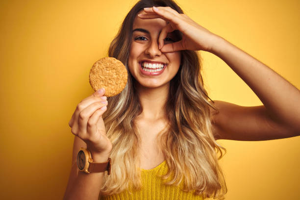 jeune belle femme mangeant le biscuit au-dessus du fond d'isolement gris avec le sourire heureux faisant le signe d'ok avec la main sur l'oeil regardant par des doigts - cookie women eating beautiful photos et images de collection