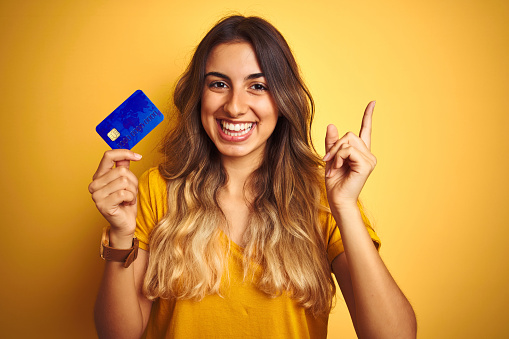 Young beautiful woman holding credit card over yellow isolated background very happy pointing with hand and finger to the side