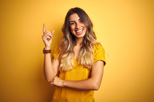 Young beautiful woman wearing t-shirt over yellow isolated background with a big smile on face, pointing with hand and finger to the side looking at the camera.