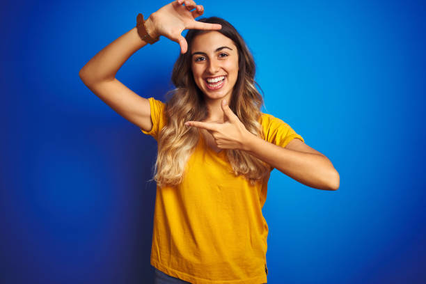 행복 한 얼굴로 손과 손가락으로 프레임을 만드는 파란색 고립 된 배경 에 노란색 티셔츠를 입고 젊은 아름다운 여자. 창의력과 사진 개념. - teenage girls ideas blue yellow 뉴스 사진 이미지