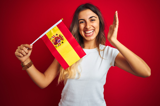 Young beautiful woman holding spanish flag over red isolated background very happy and excited, winner expression celebrating victory screaming with big smile and raised hands