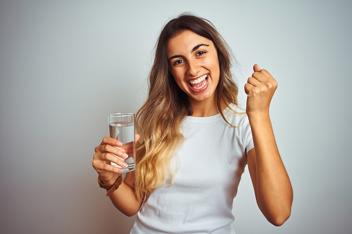 Young beautiful woman drinking a glass of water over white isolated background screaming proud and celebrating victory and success very excited, cheering emotion