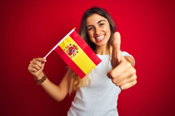 赤い孤立した背景の上にスペインの旗を持つ若い美しい女性は、okサインをやって大きな笑顔で幸せ、指で親指、優れた兆候 - interior wall 写真 ストックフォトと画像