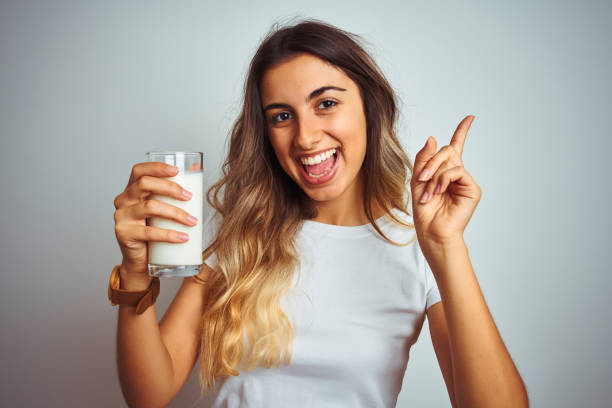 白い孤立した背景の上にミルクのグラスを飲む若い美しい女性は、側に手と指で指して非常に幸せ - food white caucasian color image ストックフォトと画像