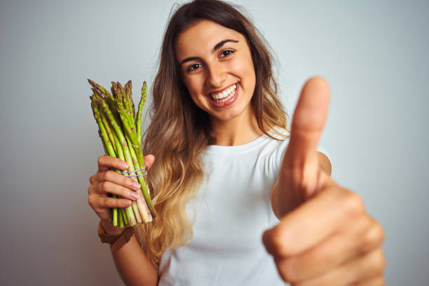 Young beautiful woman eating asparagus over grey isolated background happy with big smile doing ok sign, thumb up with fingers, excellent sign Young beautiful woman eating asparagus over grey isolated background happy with big smile doing ok sign, thumb up with fingers, excellent sign eating asparagus stock pictures, royalty-free photos & images