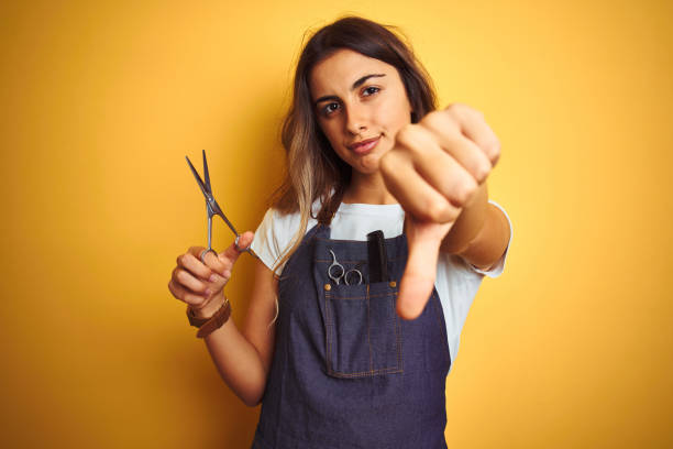Young beautiful hairdresser woman holding scissors over yellow isolated background with angry face, negative sign showing dislike with thumbs down, rejection concept Young beautiful hairdresser woman holding scissors over yellow isolated background with angry face, negative sign showing dislike with thumbs down, rejection concept angry hairstylist stock pictures, royalty-free photos & images