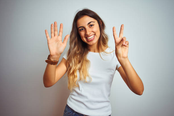 カジュアルな白いtシャツを着た若い美しい女性は、自信を持って幸せに微笑みながら、7番の指を見せ、指を上げて見せます。 - interior wall 写真 ストックフォトと画像