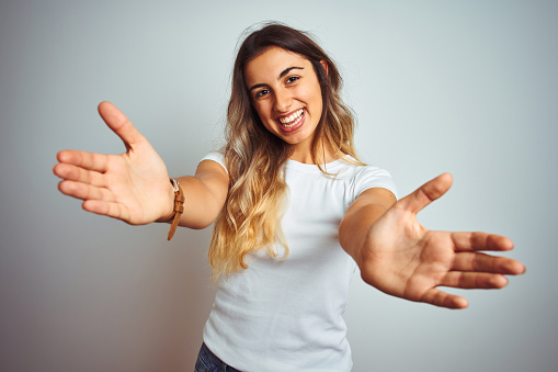 Young beautiful woman wearing casual white t-shirt over isolated background looking at the camera smiling with open arms for hug. Cheerful expression embracing happiness.