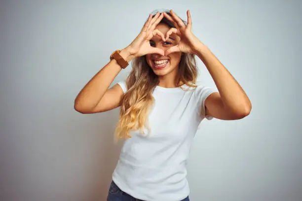 Young beautiful woman wearing casual white t-shirt over isolated background Doing heart shape with hand and fingers smiling looking through sign