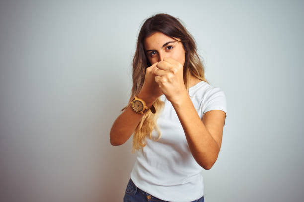 jeune belle femme utilisant le t-shirt blanc occasionnel au-dessus du fond d'isolement prêt à se battre avec le geste de défense de poing, visage fâché et bouleversé, peur du problème - ready to fight photos et images de collection