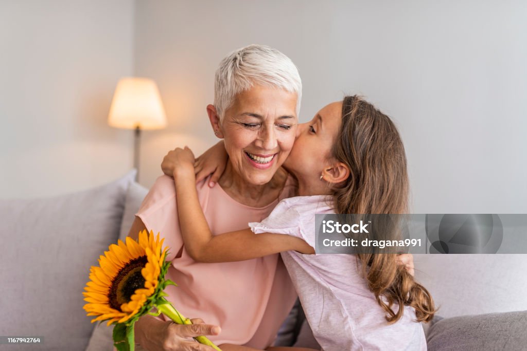 Happy little girl giving bouquet of flowers to her grandmother Happy grandmother hugging small cute grandchild thanking for flowers presented, excited granny embrace granddaughter congratulating her with birthday, making surprise presenting bouquet Grandmother Stock Photo