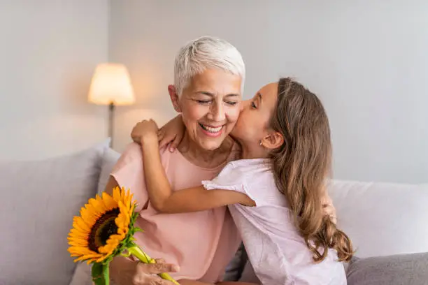 Happy grandmother hugging small cute grandchild thanking for flowers presented, excited granny embrace granddaughter congratulating her with birthday, making surprise presenting bouquet