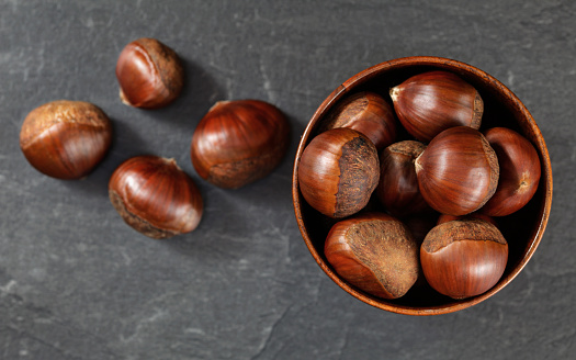 Small wooden cup with chestnuts, some scattered on black slate like board.