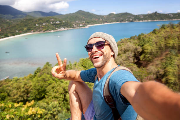 Young male hipster traveler doing selfie overlooking the tropical sea. Adventure, vacation, wonderlust, internet, technology concept. stock photo