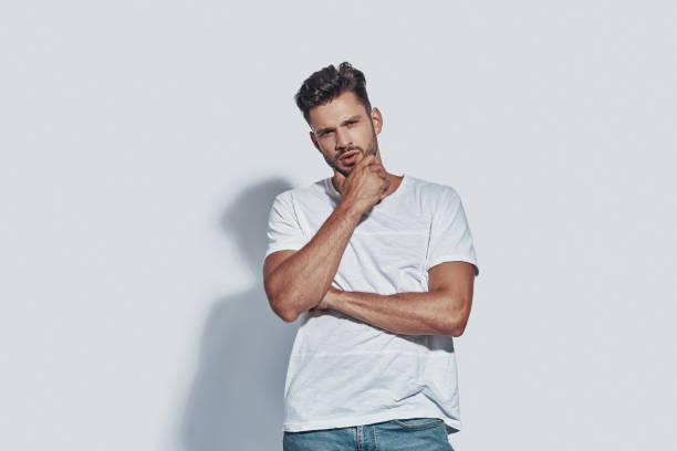 istock Handsome young man looking at camera and keeping hand on chin while standing against grey background 1167953802