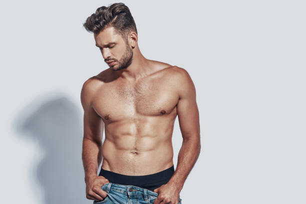 handsome young shirtless man taking off his jeans while standing against grey background - shirtless imagens e fotografias de stock