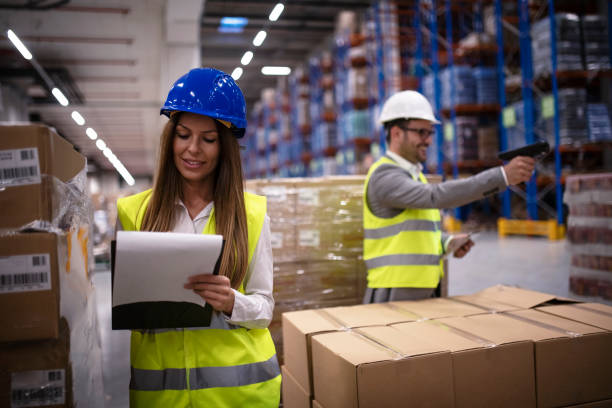 trabalhador fêmea da fábrica no uniforme reflexivo com capacete do capacete de segurança que verific a chegada nova dos bens no armazém quando o trabalhador que usa o leitor de código de barra no fundo. logística e distribuição. - bar code reader bar code checklist box - fotografias e filmes do acervo