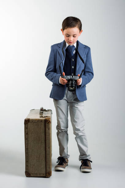 8 year old boy dressed up in suit, standing next to vintage suitcase, looking at photo camera taking up a vintage suitcase. travel concept
