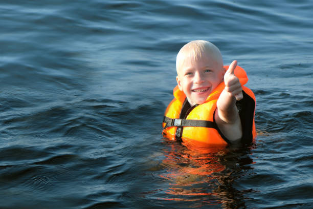 blond little boy in orange life vest swimming in sea. boy shows thumb up stock photo
