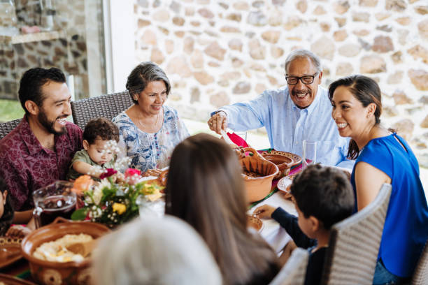 latin senior mann serviert das essen zu seiner familie am esstisch - familie mit mehreren generationen stock-fotos und bilder
