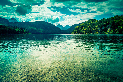 Wide angle color image depicting the tranquil scene of a crystal clear still blue mountain lake. In the far distance is a dramatic mountain range with cloudscape. Room for copy space.