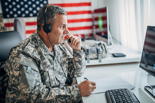 Army soldier wearing headphones ,using computer, and writing notes