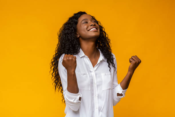 Black girl smiling and raising clenched fists in the air Emotional african american woman smiling and raising clenched fists in the air, feeling excited, yellow studio background raised fist photos stock pictures, royalty-free photos & images