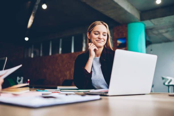 concepteur grapic féminin réussi regardant le tutoriel au sujet des idées créatrices à l'ordinateur portable pendant le processus de travail dans le bureau. étudiant positif avec le cheveu blond lisant des nouvelles d'affaires sur le netbook - conférence en ligne photos et images de collection