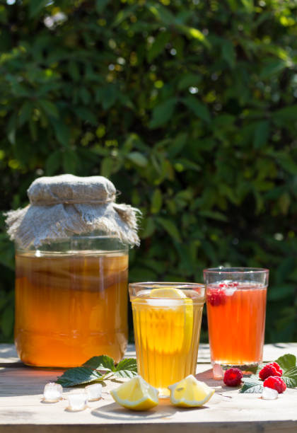 istock Fermented Kombucha drink in a glass jar and two glasses with a drink with lemon and raspberry berry and ice, in the foreground, on a Sunny summer day in the garden on a wooden table. 1168029850