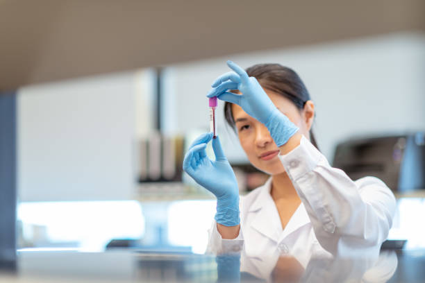 Biochemist analyzing small sample of blood A female chemist of Chinese descent is working solo in scientific lab. She is standing at a work station. The woman is holding up a small vial of blood and is analyzing the medical sample. She is wearing a lab coat and protective gloves. medical sample stock pictures, royalty-free photos & images