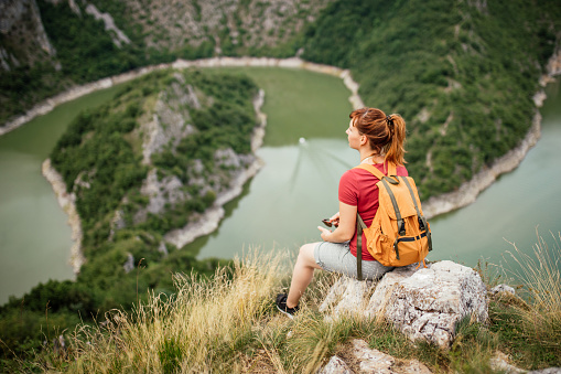 Young hiker woman enjoying the view from the top of mountain over beautiful river, looking through binoculars