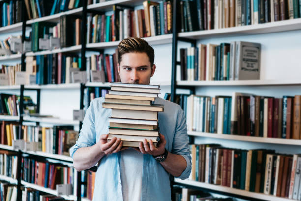 Overworked persistent young man in casual wear holding many literature books in hands for science course work.Intelligent student with textbooks standing in library interior with book shelves joven persistente sobrecargado de trabajo en ropa casual sosteniendo muchos libros de literatura en las manos para el trabajo del curso de ciencias. estudiante inteligente con libros de texto de pie en el interior de la biblioteca con estantes de libros