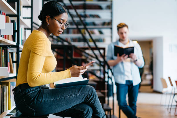 istock Pensive african american young woman reading incoming sms message on smartphone while preparing for exam in library.Students spending leisure time in university bookstore reading literature books 1168056903