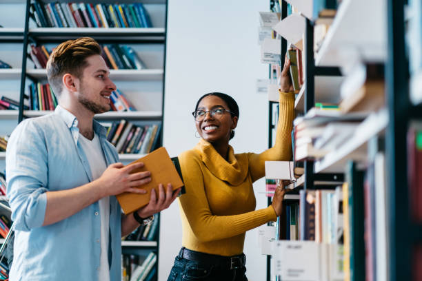 istock Positive two multicultural young people teamworking in library.Cheerful diverse hipster students dressed in formal wear pulling books from shelf choosing literature for writing course work 1168056917
