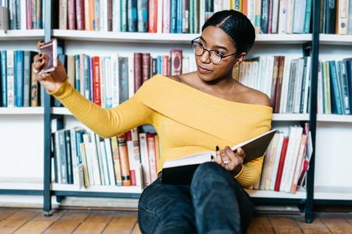 African american young woman student in eyewear making selfie photo on front camera of smartphone for sharing in networks sitting with literature book on floor and preparing for exam in library