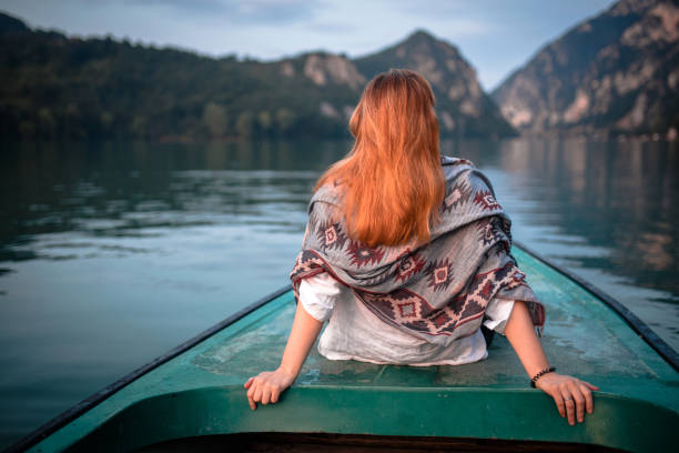 Young woman enjoys dawn on boat on lake, looking away in mountains