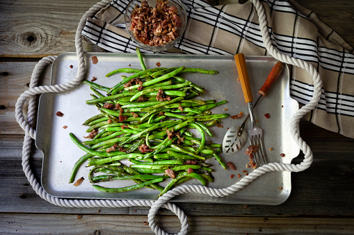 horizontal vintage look of stir fried green beans in a metal cooking sheet sprinkled with real bacon bits on a rustic wood table